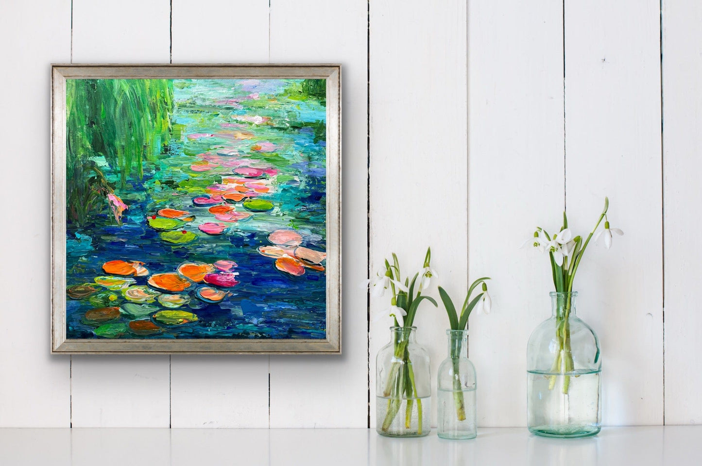 Framed impressionist painting by Maria-Victoria Checa of water lilies on a white wall with three glass bottles containing plants below.