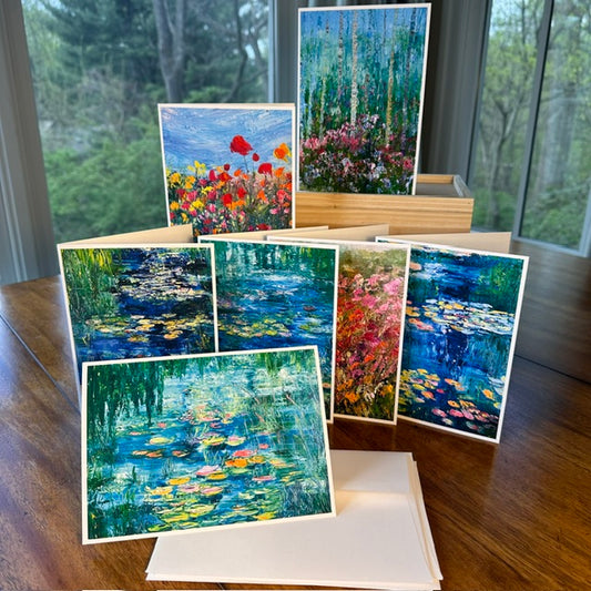Seven impressionist art greeting cards fanned out on a wood table — water lily pond, wildflower field, birch forest, and flower meadow designs by Washington DC artist Maria-Victoria Checa