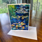 Dark blue water lily pond greeting card with vibrant orange and pink lilies and green reeds — impressionist art card by Maria-Victoria Checa