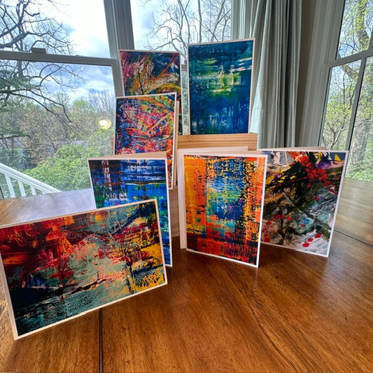 Seven bold abstract art greeting cards fanned out on a wood table showing vibrant colors in blue, orange, red and multicolor — Checa Art Bleu collection card set by Washington DC artist Maria-Victoria Checa