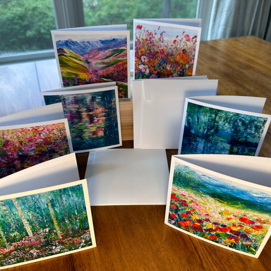 Seven square impressionist art greeting cards fanned out on a wood table showing mountain valley, flower fields, forest and water lily pond designs — Checa Art Sunset Lilies card set by Washington DC artist Maria-Victoria Checa