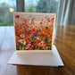 Wildflower field greeting card with luminous red, orange, pink and blue blooms under a soft sky — square impressionist art card by Maria-Victoria Checa