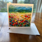 Poppy field greeting card with vibrant red and yellow flowers stretching toward rolling green hills and blue sky — square impressionist art card by Maria-Victoria Checa