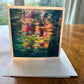 Water lily pond greeting card with green lily pads and warm pink and orange sunset reflections on dark teal water — square impressionist art card by Maria-Victoria Checa