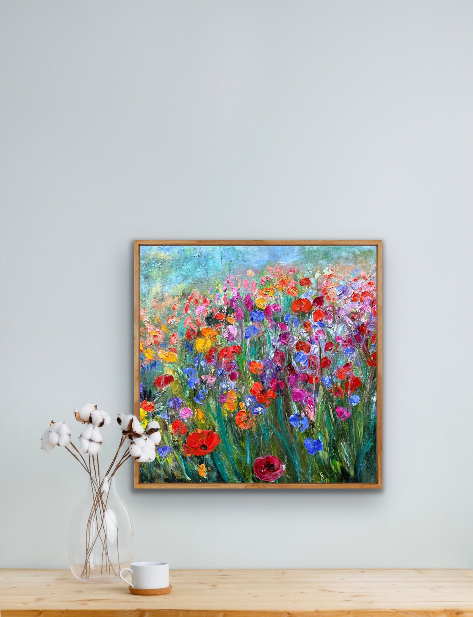 Framed painting of colorful flowers on a wall above a wooden surface with a vase and cup.