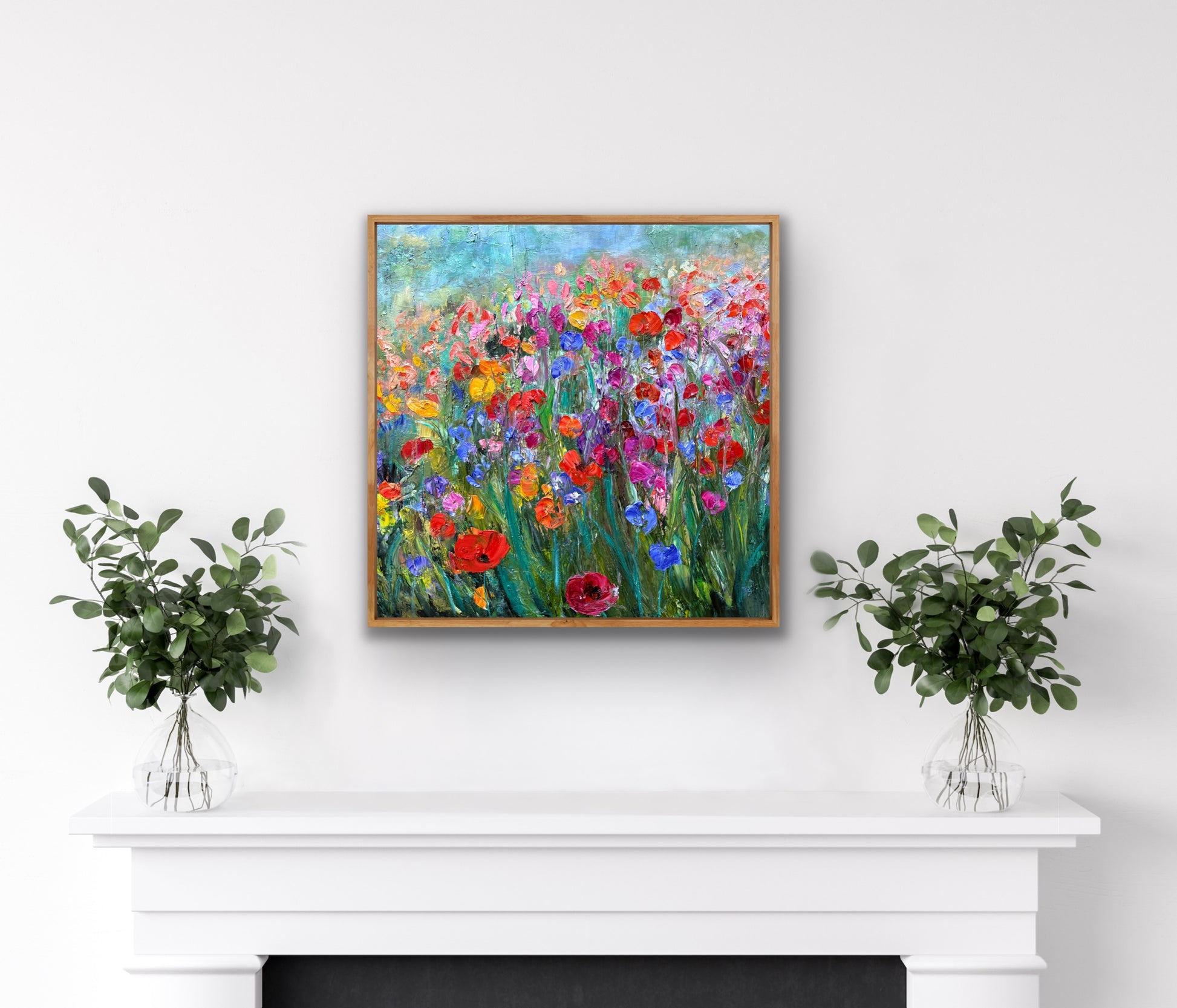 Framed painting of a colorful floral scene on a white wall above a fireplace with two small potted plants.