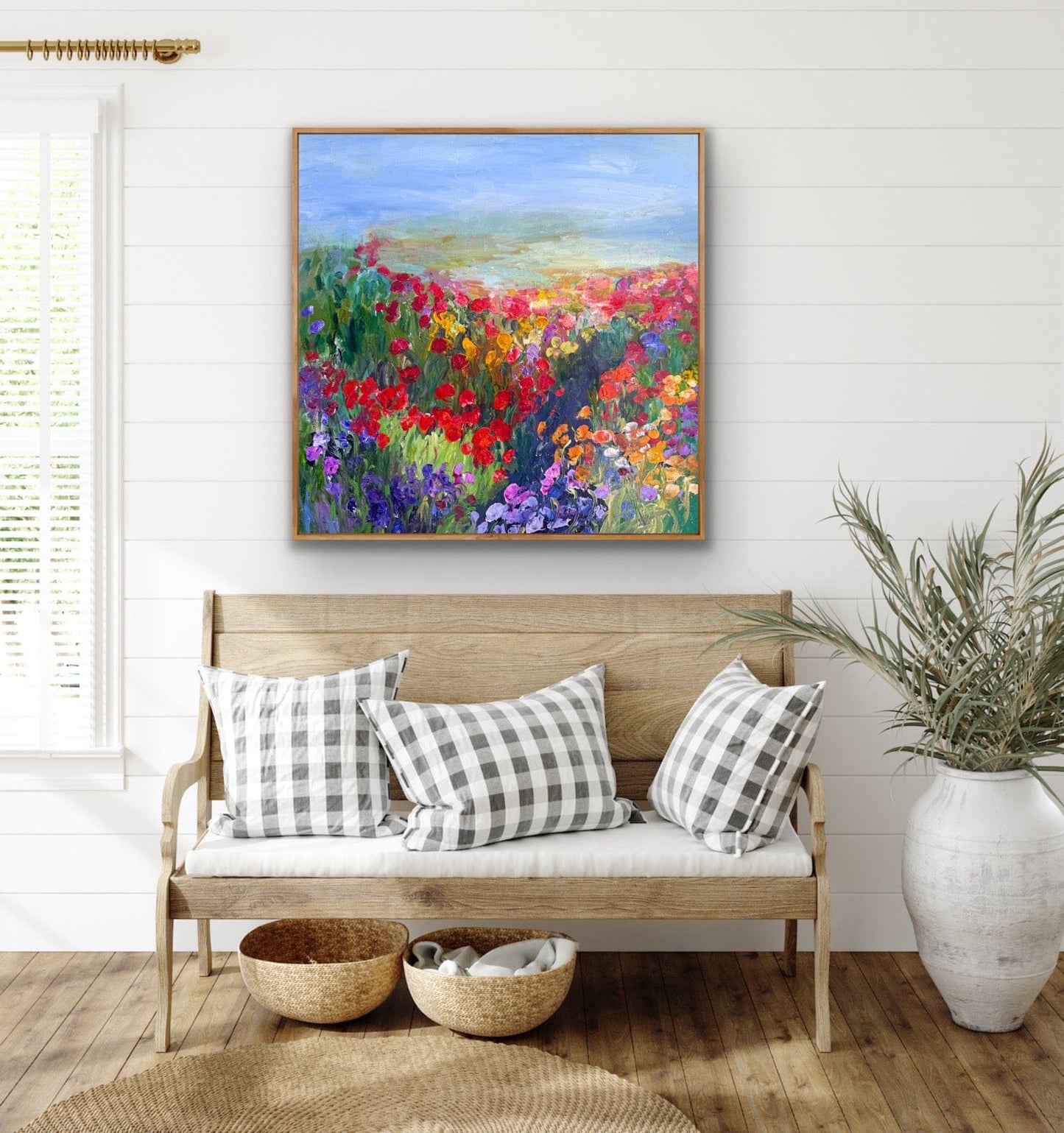 A colorful impressionist flower landscape painting titled “Path in the Meadow” by Maria-Victoria Checa displayed above a light wood farmhouse bench with gingham pillows in a bright, coastal-style interior.