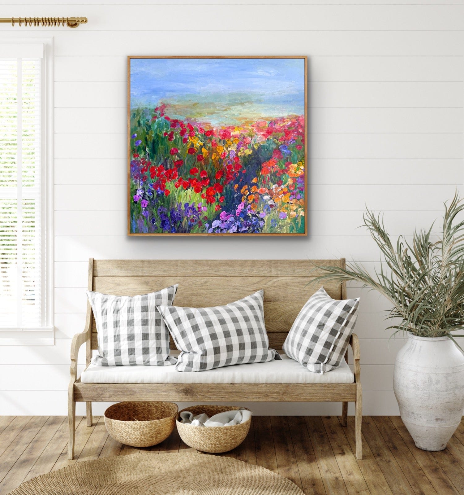 A colorful impressionist flower landscape painting titled “Path in the Meadow” by Maria-Victoria Checa displayed above a light wood farmhouse bench with gingham pillows in a bright, coastal-style interior.