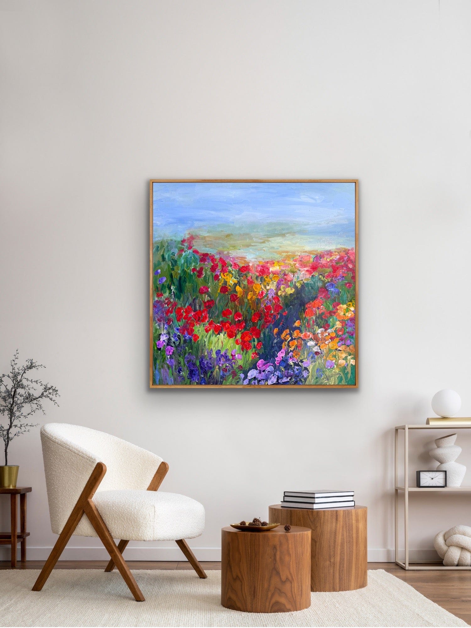 A framed impressionist flower-path painting titled “Path in the Meadow” by Maria-Victoria Checa displayed in a modern neutral living room above a white armchair and wooden side tables.