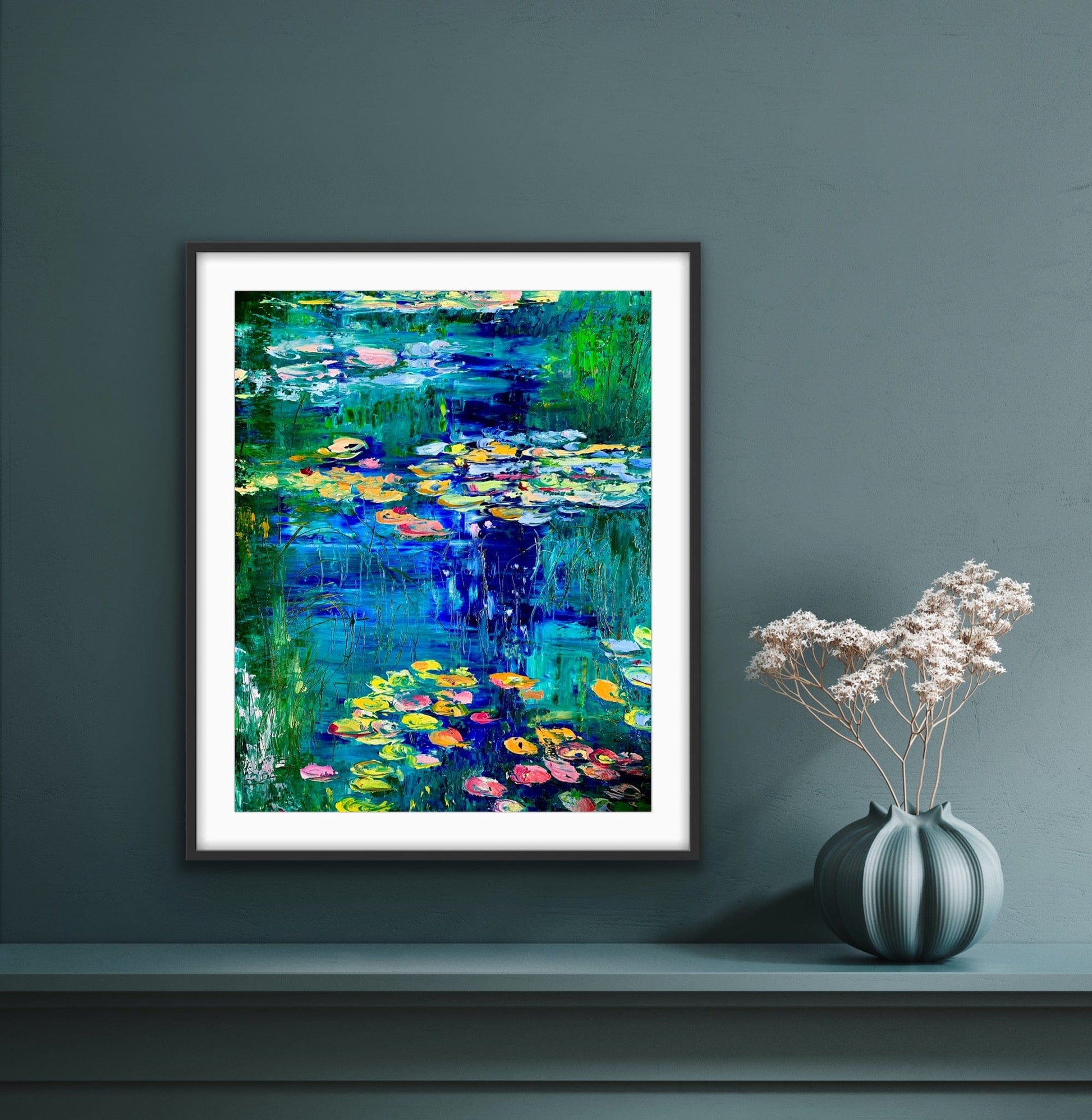 Framed paper print with mat of “Water Lily Pond” by Maria-Victoria Checa displayed on a blue wall above a console, showing vibrant water lilies and reflective blue water.